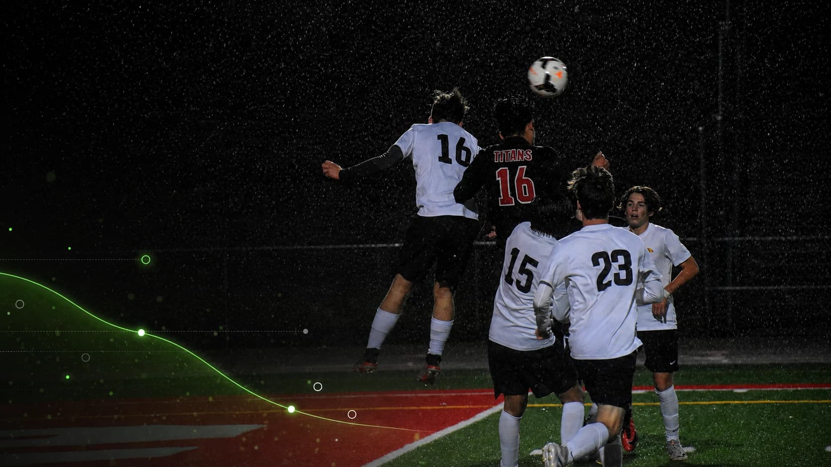Footballers contest a header during a night match in heavy rain.