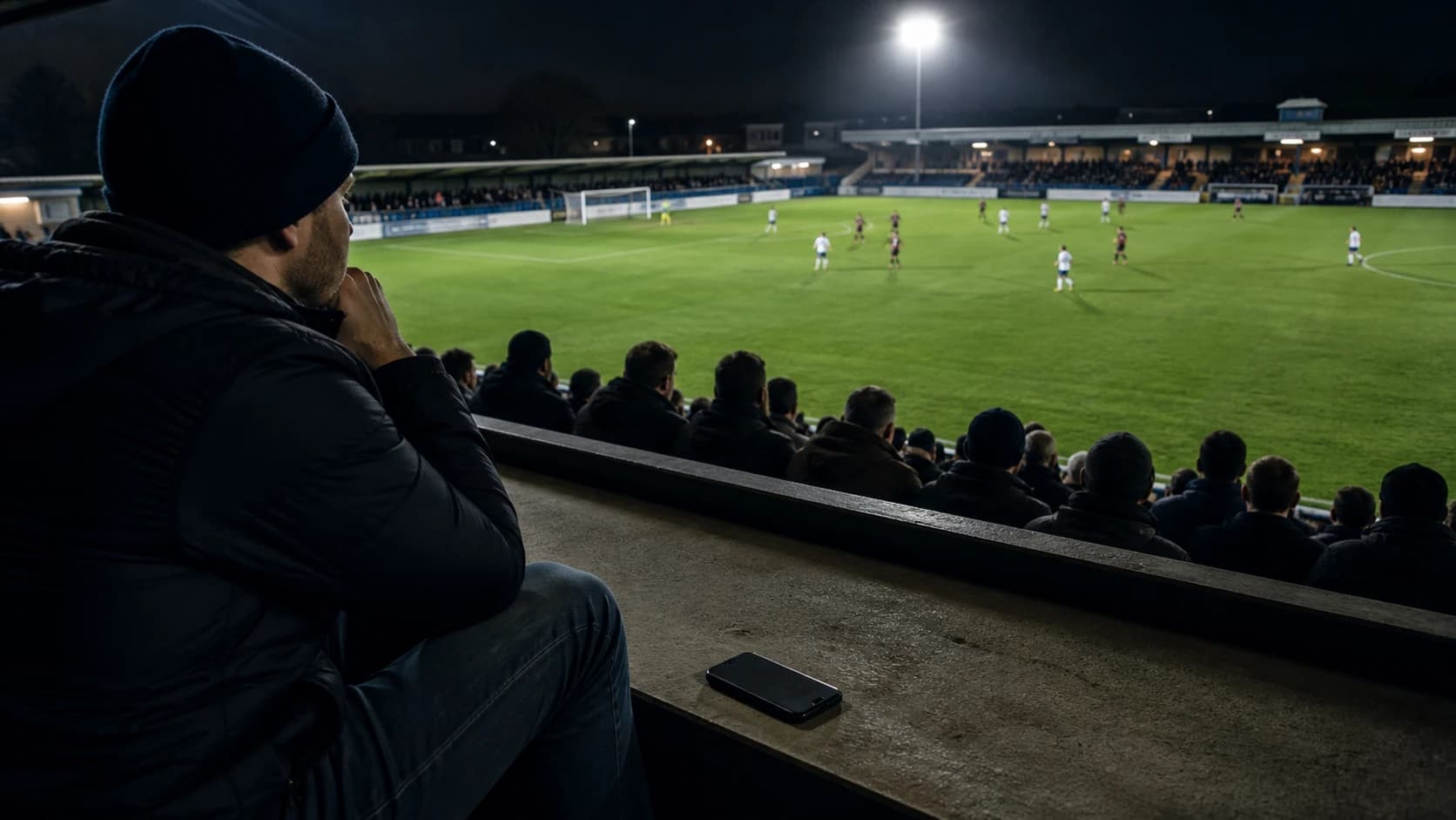 An adult spectator watches a floodlit football match continue with an unbranded phone placed face down beside him.